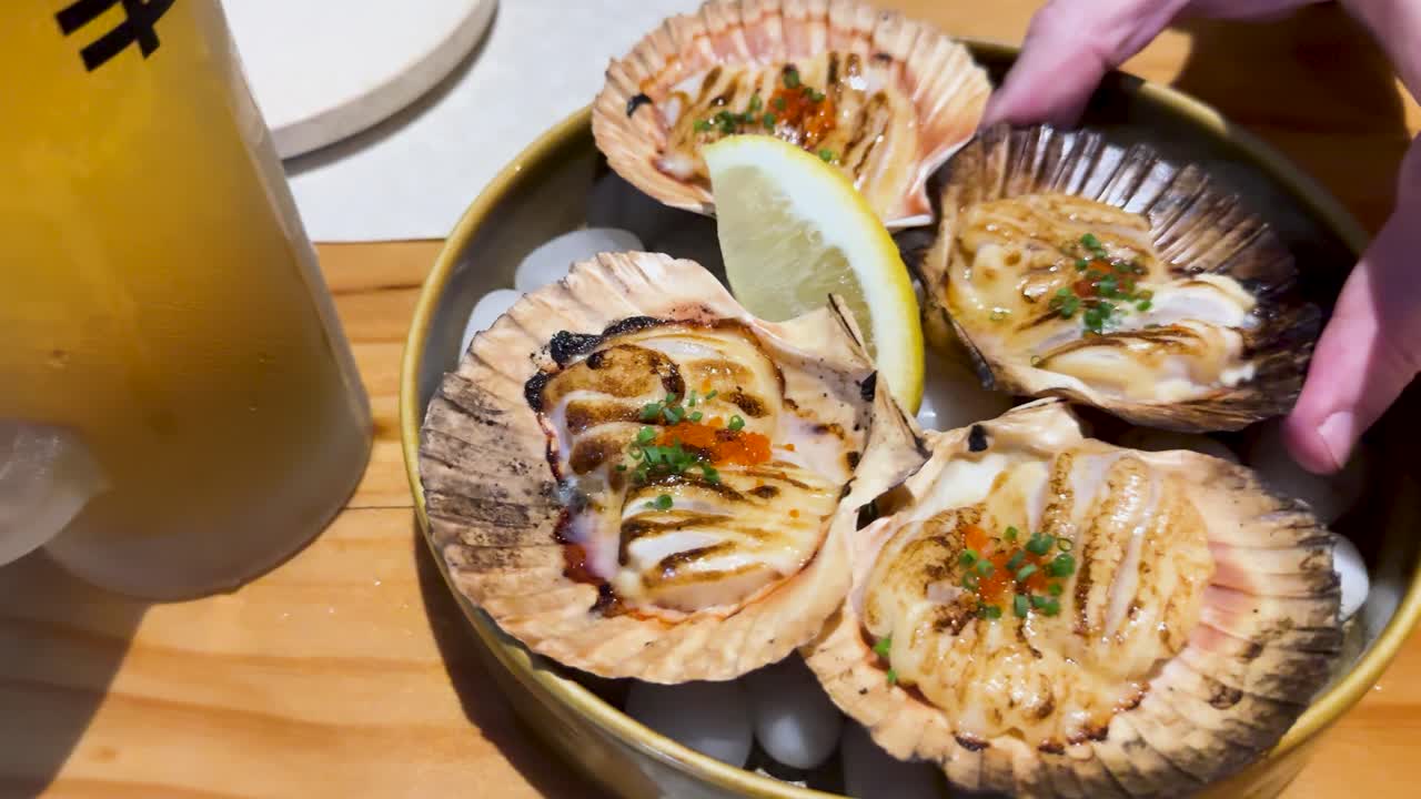 Fried scallops served in shells with lemon garnish, accompanied by a drink. Bright lighting highlights the dish's texture