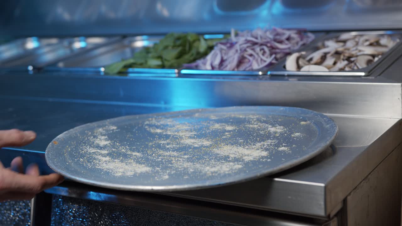 Et nærbilde av hendene til en dyktig kokk som legger et pizzabrett på en stålkjøkkenbenk mens kameraet panorerer med bevegelsene hans.