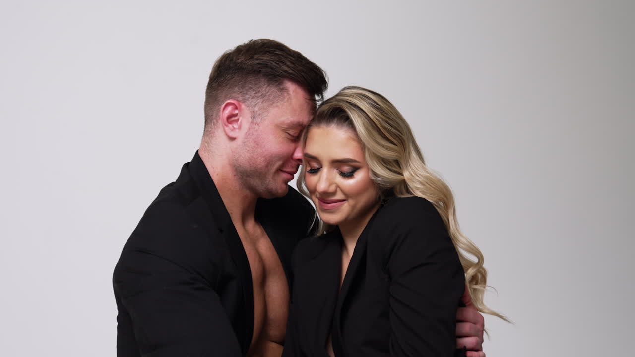 Beautiful Caucasian couple wearing black jackets sit close to each other. Man and woman in love embracing. Studio footage. White backdrop.