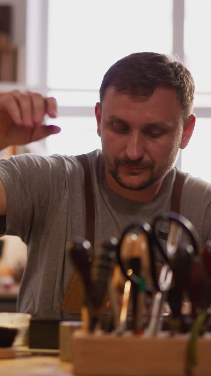 Skilled leathersmith turns on table lamp processing leather in workshop closeup. Bearded man does craft item in small studio. Master at workplace