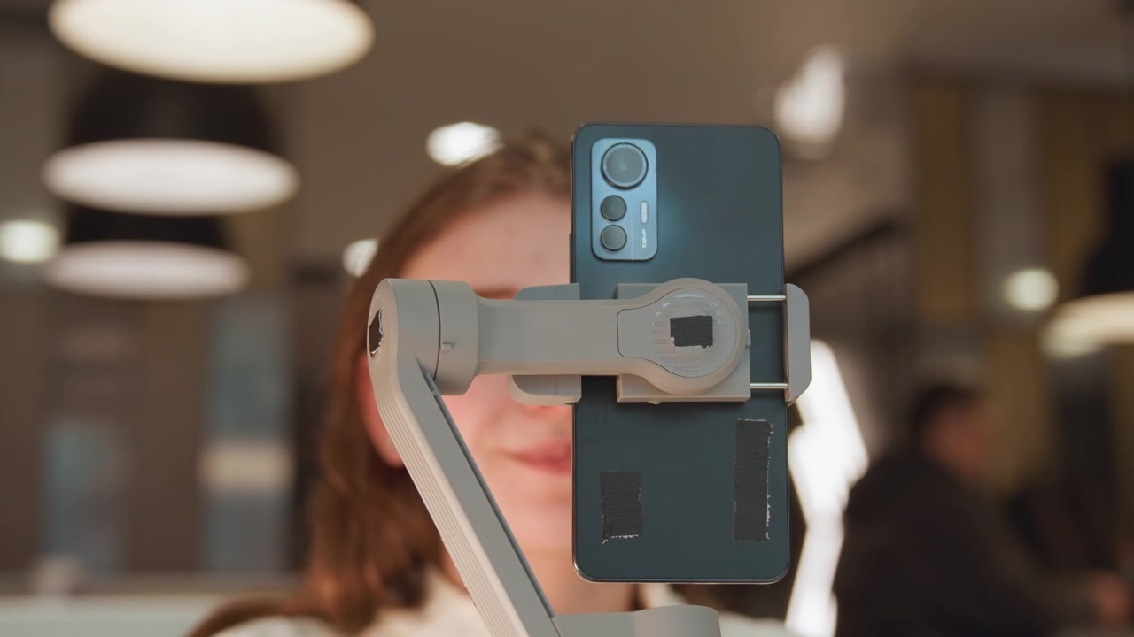 Jovem mulher sentada em um espaço de trabalho interno aconchegante sorri com os olhos fechados enquanto um smartphone em um braço ajustável está diretamente na frente do seu rosto, capturando uma reação calma durante criação e interação social relaxadas.