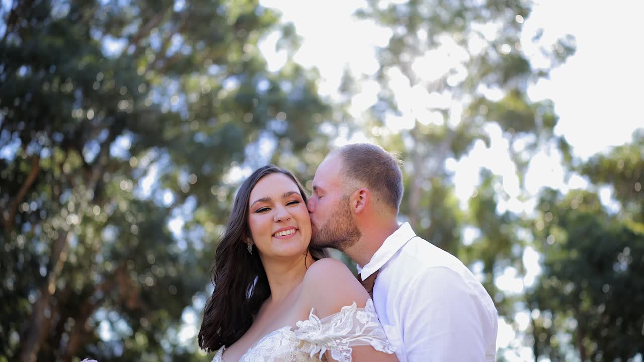 Joven novio besa a la novia sonriente durante las fotos en un soleado jardín trasero