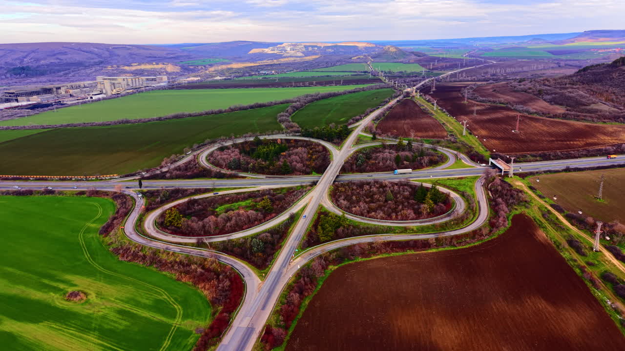 Vejkryds omgivet af grønne marker med et industrianlæg i baggrunden. Luftfoto af motorvejskløverbladsanlæg nær fabrik.
