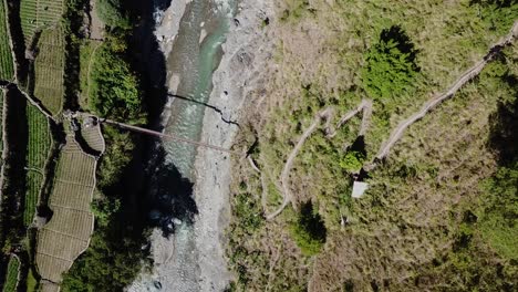 Top-down-descending-on-mountainous-zig-zag-dirt-gravel-trail-leading-to-metal-suspension-bridge-river-crossing-emerald-mountain-water-grey-rocks-green-trees