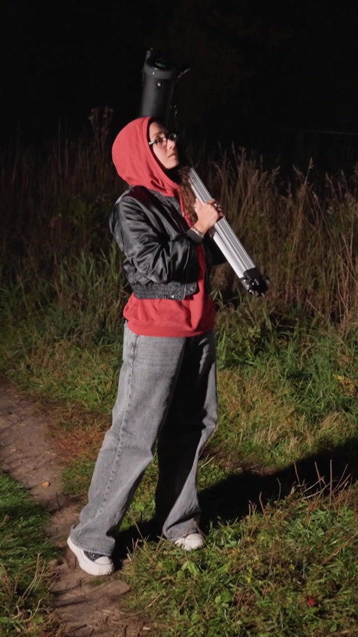 Individual adjusts telescope on grassy trail surrounded. Subject holds scope horizontally and stands upright. Outdoor setting features narrow dirt trail with grassy edges