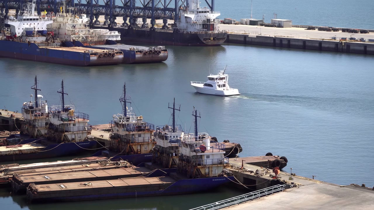Una pequeña lancha motora blanca se mueve sobre aguas tranquilas y azules del puerto junto a barcos de pesca amarrados, con edificios portuarios industriales, grúas y estructuras de muelle visibles