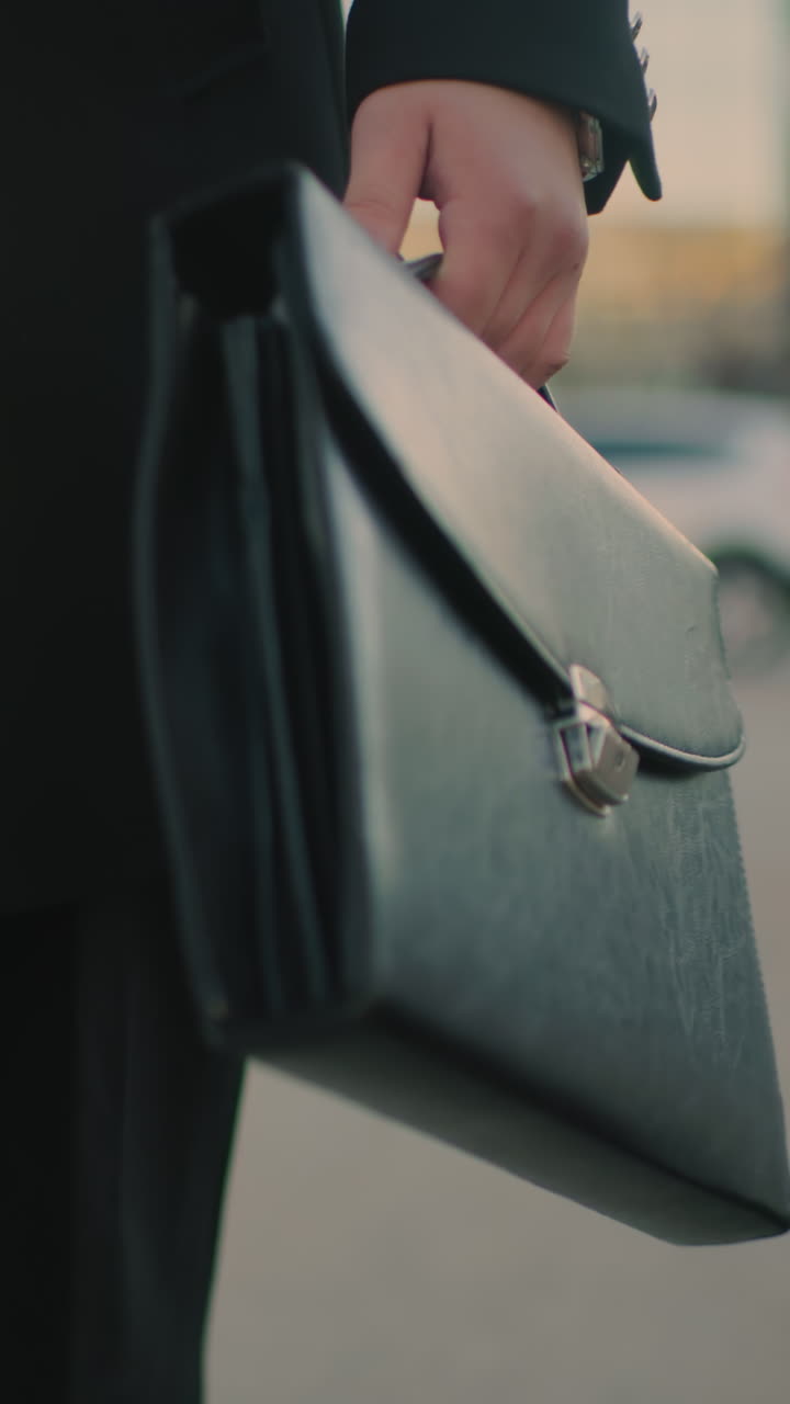 Close-up of man in black outfit walking outdoors with briefcase in left hand and coffee in right hand, blurred background features parked cars