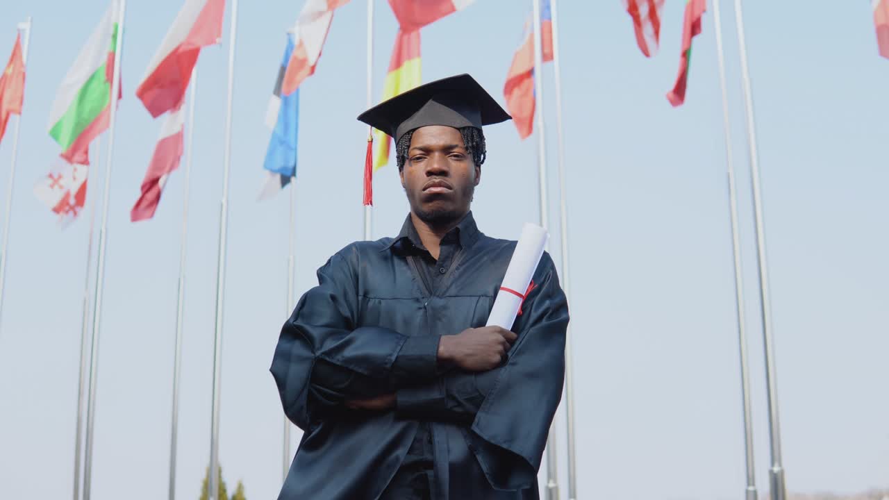 Young african american male graduate standing in front of the camera with a diploma in his hands. The student has a square master's hat and stands outside with the international flags on background.