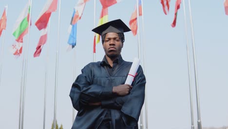 joven graduado afroamericano de sexo masculino de pie frente a la cámara con un diploma en las manos. el estudiante tiene un sombrero de maestro cuadrado y está de pie afuera con las banderas internacionales en el fondo.