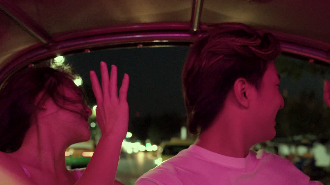 Una pareja sonriente monta en un tuk tuk abierto de noche, con luces de neón de la ciudad y movimiento de cámara en mano