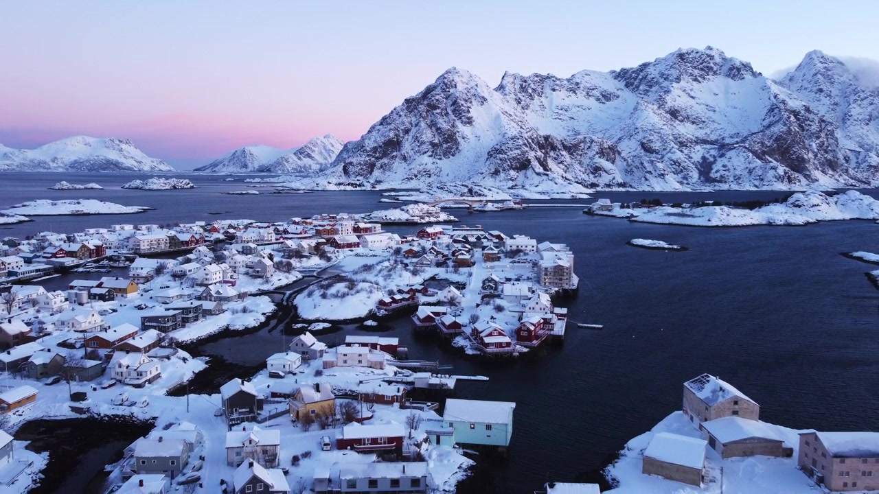 Luftperspektiv med drone som flyr over Henningsvær fiskevær dekket av snø ved daggry, Lofoten, Norge. Historisk nordisk arkitektur og vinterlig kystlandskap ved soloppgang