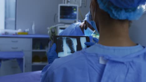 Cirujanos-De-Raza-Mixta-Con-Ropa-Protectora-Mirando-Radiografías-De-Pulmón-En-Una-Tableta-En-El-Quirófano