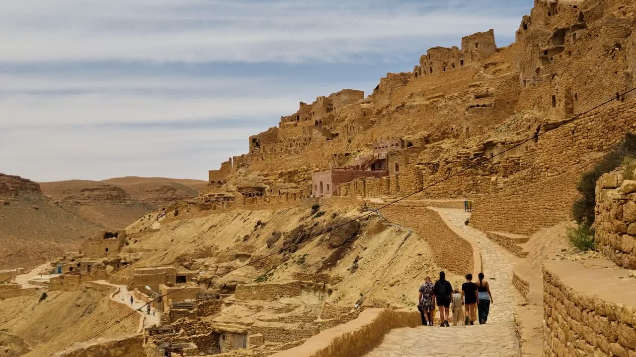 Tourists slowly walking down a path in a historic Berber town with medieval buildings and a fortress in the background. Filmed in old Chenini, Tataouine region of Tunisia on a pleasant spring day