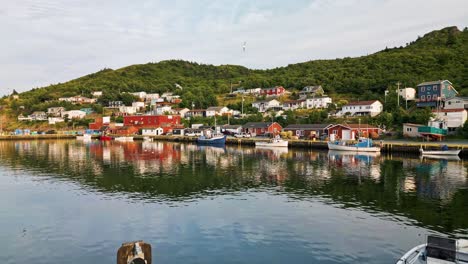 a drone glides over a calm newfoundland fishing village, revealing colorful houses on a green hillside, wooden docks and boats, and reflections shimmering on the bay