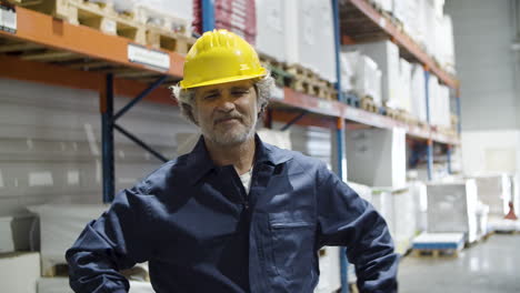 trabajador senior feliz en casco de pie en el almacén y mirando a la cámara con los brazos cruzados