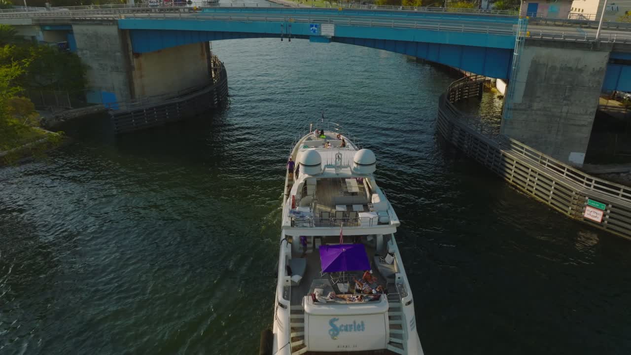 Forwards tracking of luxury yacht passing under road bridge. People enjoying sunny afternoon on boat deck floating on river leading through city. Miami, USA