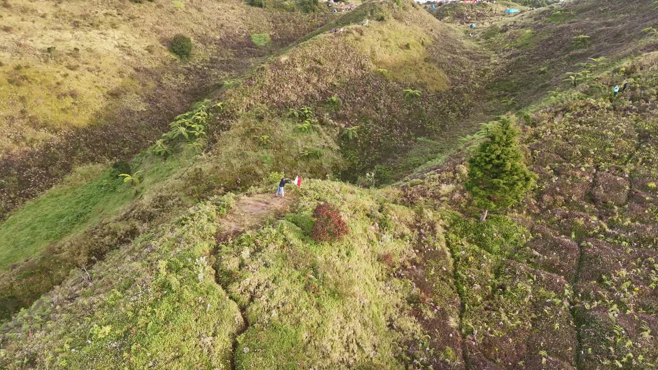 Aerial view of a man standing on a ridge waving Indonesia red-white flag surrounded by lush green hills and a natural valley basin