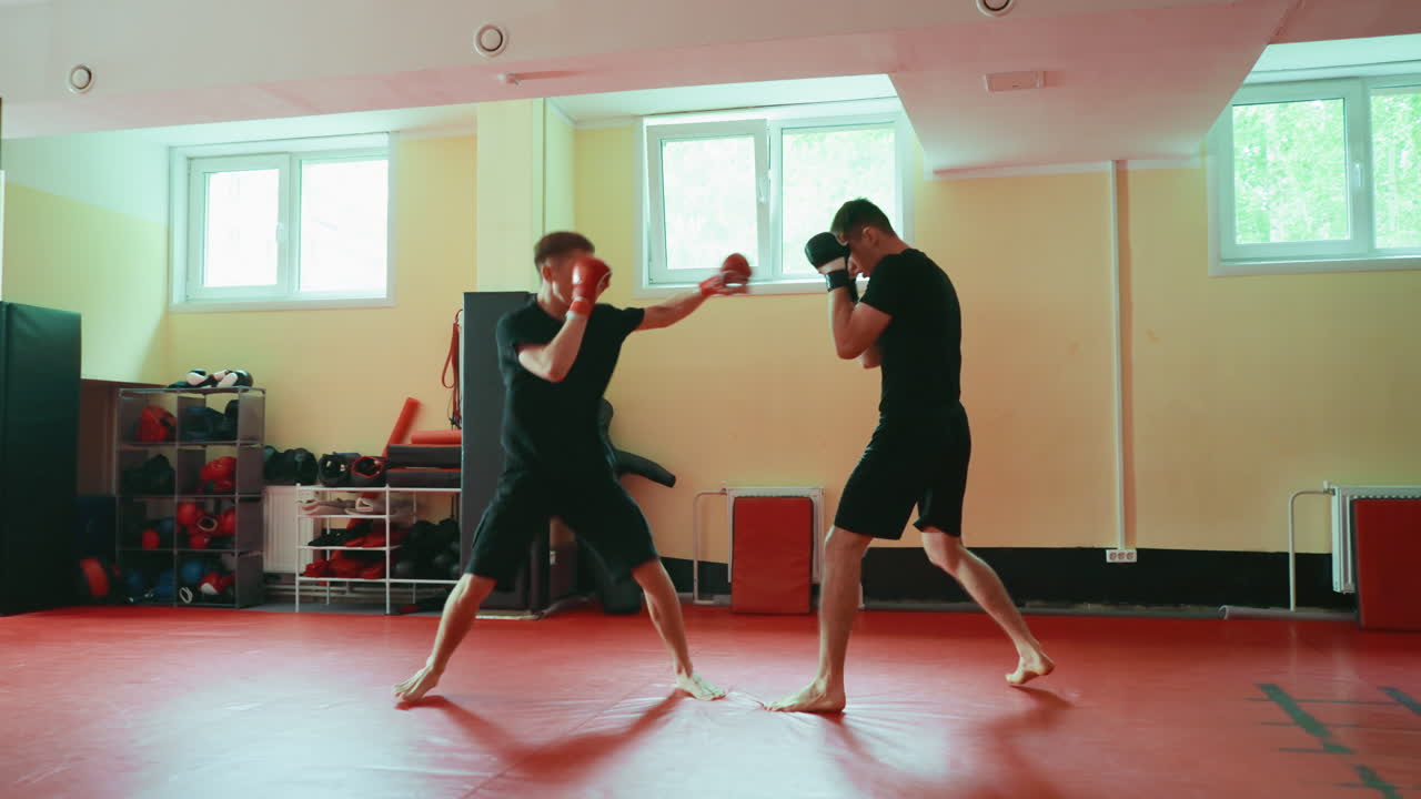 Two men training martial arts in gym, practicing sparring techniques with gloves on red mat, focused on combat movement, defensive stance, energy, and athletic performance under bright window light