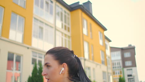 A-young-female-athlete-with-headphones-performs-jumping-exercises-in-a-city-park-against-the-background-of-buildings.-Jump-on-a-bench-in-the-park