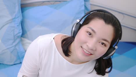 asian woman smiling using the bluetooth headphone for listening to music and looking at the camera so lovely lie in bed