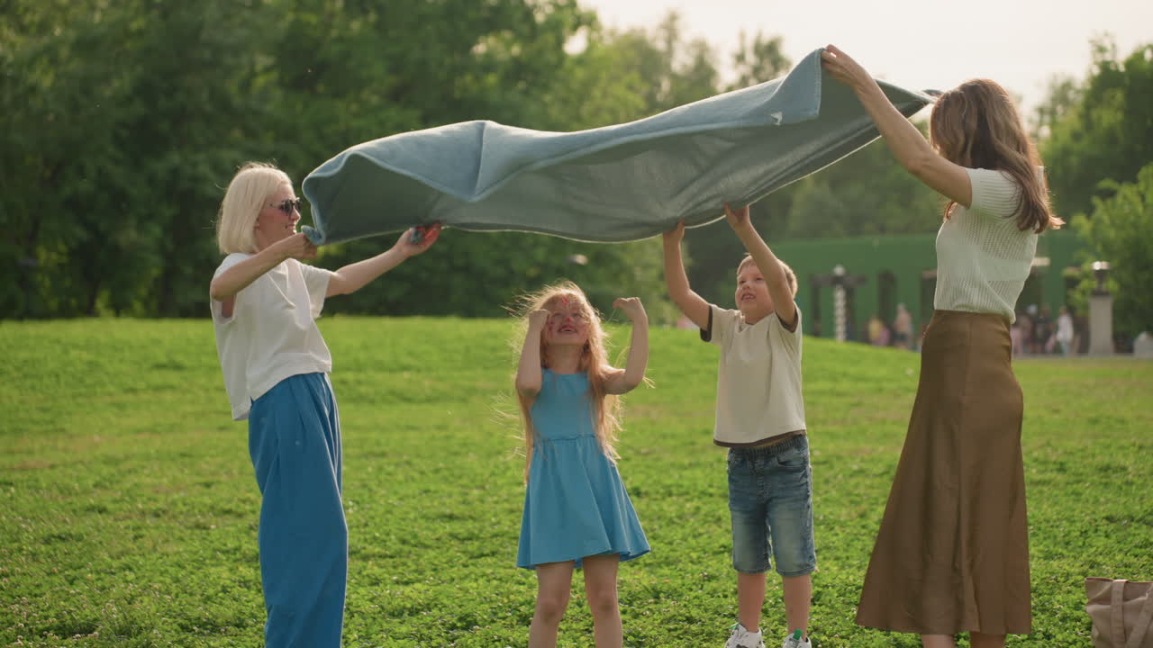 Mothers and child with girl in middle playfully lifting and swaying large mat over head in sunny park, laughter and movement, green grass background, casual summer clothing, family bonding moment