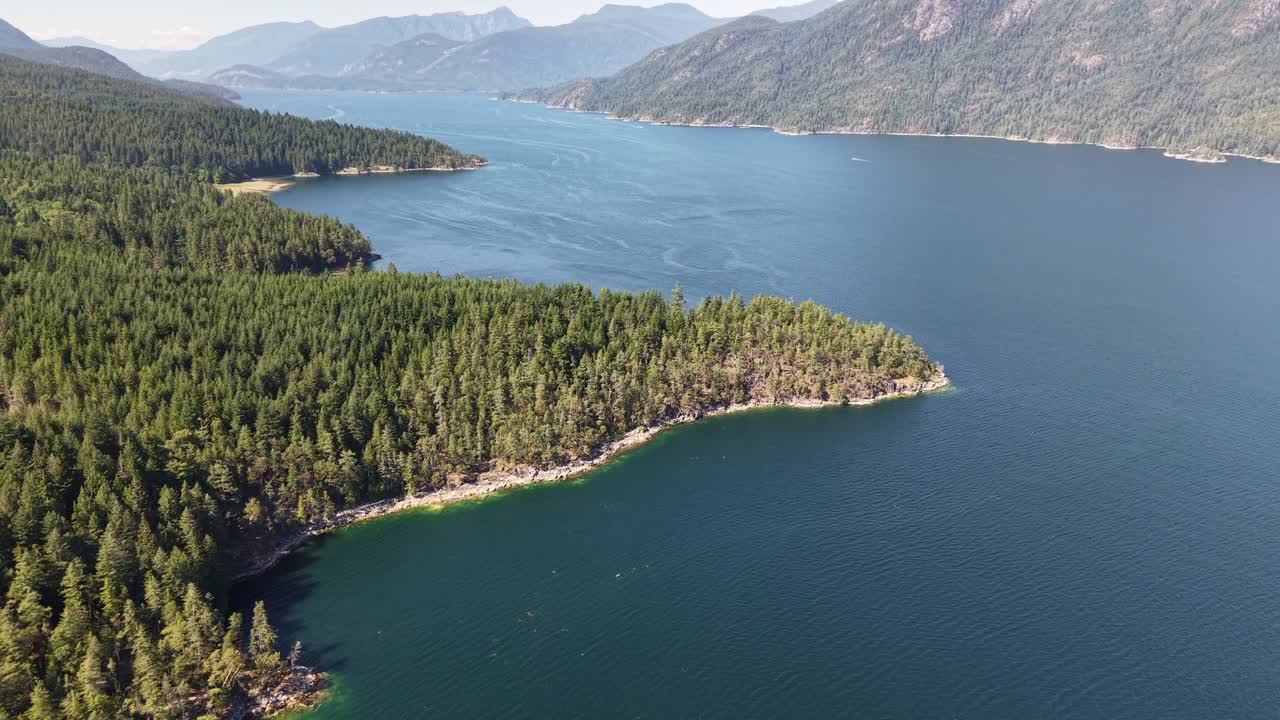 A panning shot that shows a tree covered rocky point that meets the water. Behind the point are many more rocky points, vast trees and mountains