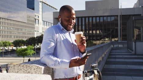 Hombre-Afroamericano-En-La-Ciudad-Con-Bicicleta-Parada-Al-Sol,-Tomando-Café,-Usando-Un-Teléfono-Inteligente