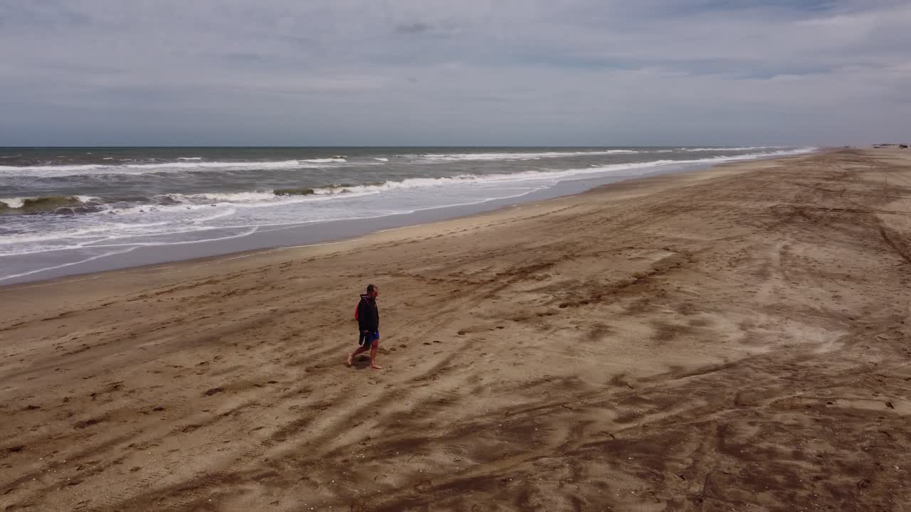 toma aérea de un hombre caminando en una playa de arena con olas del océano en el fondo durante la luz del sol - mar de las pampas, argentina