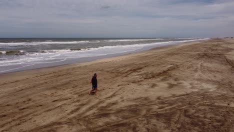 Aerial-tracking-shot-of-man-walking-on-sandy-beach-with-waves-of-ocean-in-background-during-sunlight---Mar-de-las-Pampas,Argentina