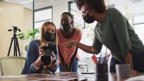 Three-diverse-female-creative-colleagues-wearing-face-masks-discussing-in-office,-looking-at-camera