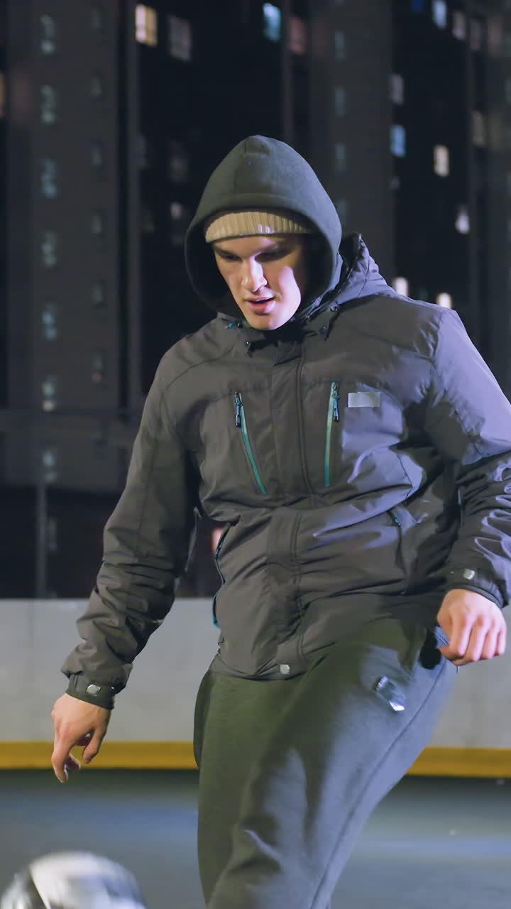 Footballer in hoodie joggling football during nighttime practice on urban sports field surrounded by high-rise buildings and bright streetlights