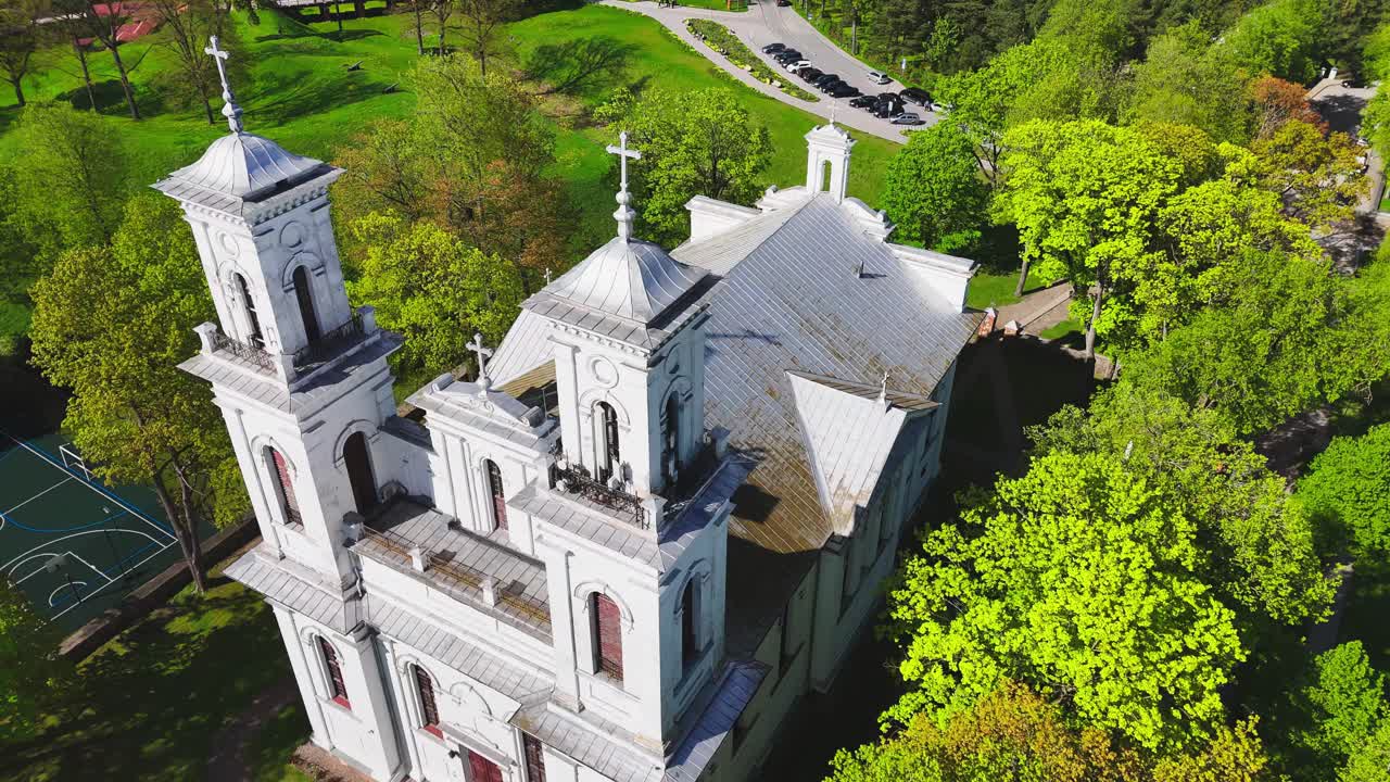 A white twin-towered church rises from a canopy of green trees in a European park, bathed in morning light and framed by spring foliage and nearby walking paths.
