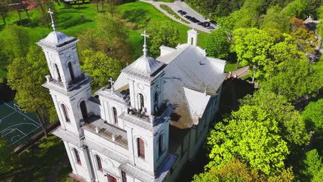a white twin-towered church rises from a canopy of green trees in a european park, bathed in morning light and framed by spring foliage and nearby walking paths.