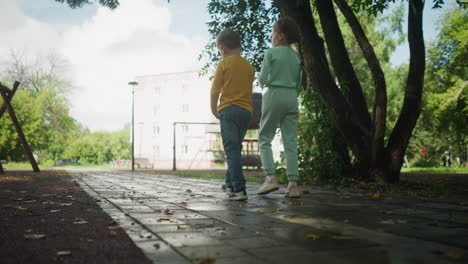 rear view of young boy and girl walking on paved path under trees toward swing set and residential building in lush urban park on sunny day, showcasing childhood friendship and outdoor exploration