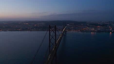 Aerial-night-orbit-of-Ponte-25-de-Abril-red-bridge-with-car-traffic-and-lights-off-the-coast-of-Lisbon-city-center