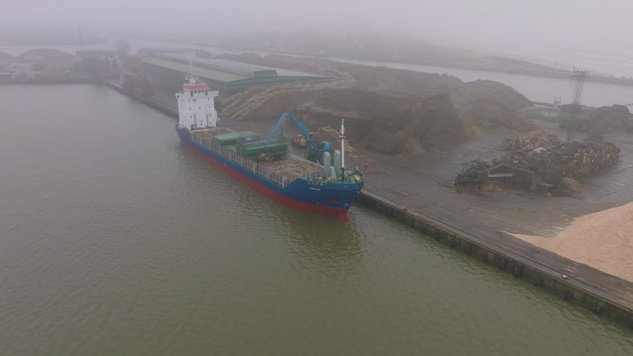 vista aérea de establecimiento de la grúa terminal de madera cargando madera en el buque de carga, puerto de liepaja, exportación de troncos de madera, día nublado con niebla y niebla, dron avanzando, inclinado hacia abajo