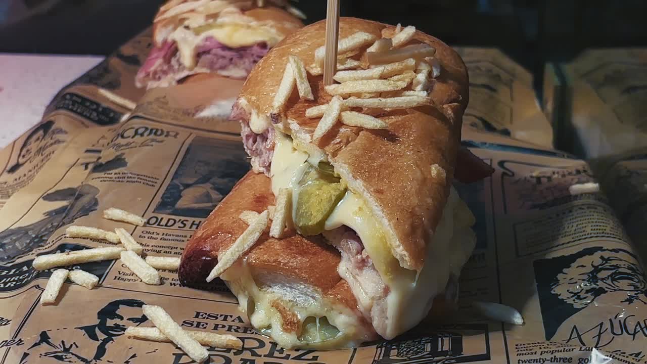 Toasted sandwich filled with melted cheese, sliced meat, and pickles topped with potato sticks and served on printed deli paper in Miami