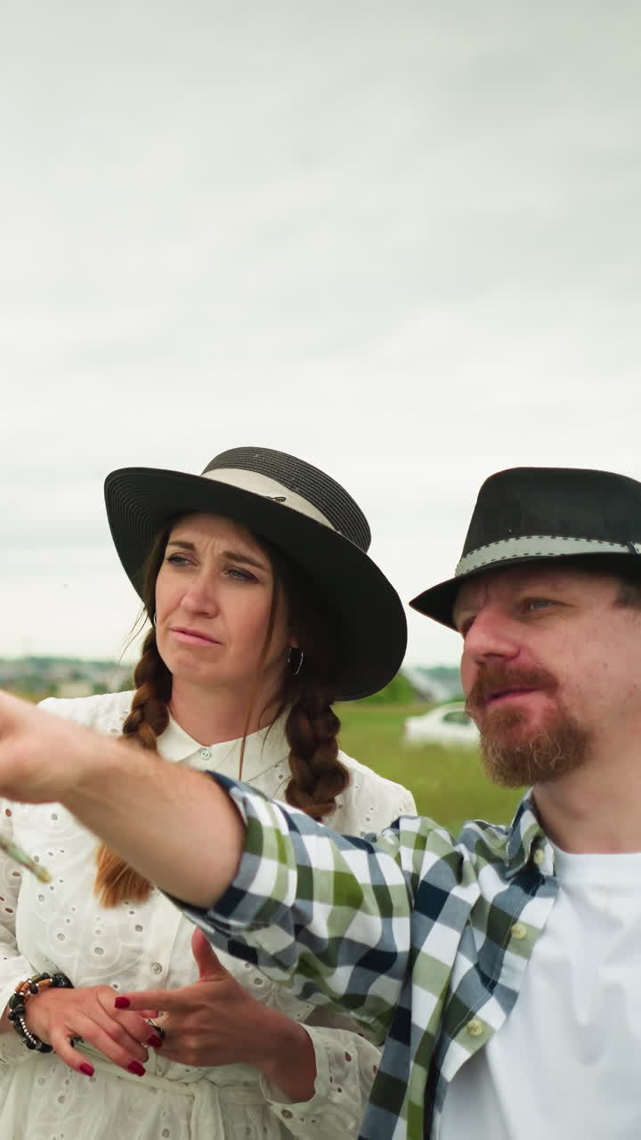 el pintor con una camisa a cuadros y un sombrero está señalando con un pincel a una mujer a su lado, vestida con un vestido blanco y sombrero. están en un campo de hierba con un lienzo parcialmente pintado en el fondo