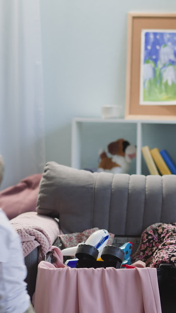 Little son puts toys into open suitcase while mother smiles looking at son and folds clothes in living room. Lady with boy prepares for vacation