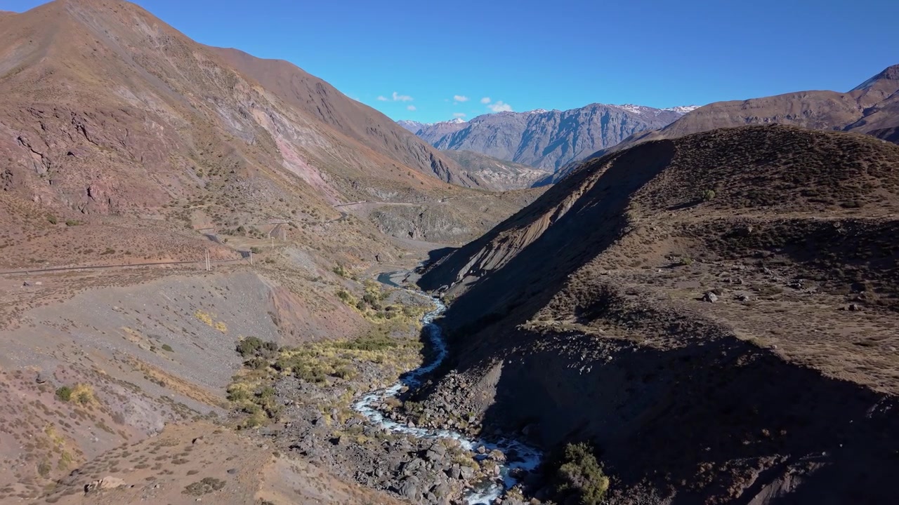 Volo in avanti che segue un impetuoso fiume di montagna cristallino che si snoda attraverso un'aspra valle rocciosa con rada vegetazione verde e lontane cime andine innevate all'orizzonte.
