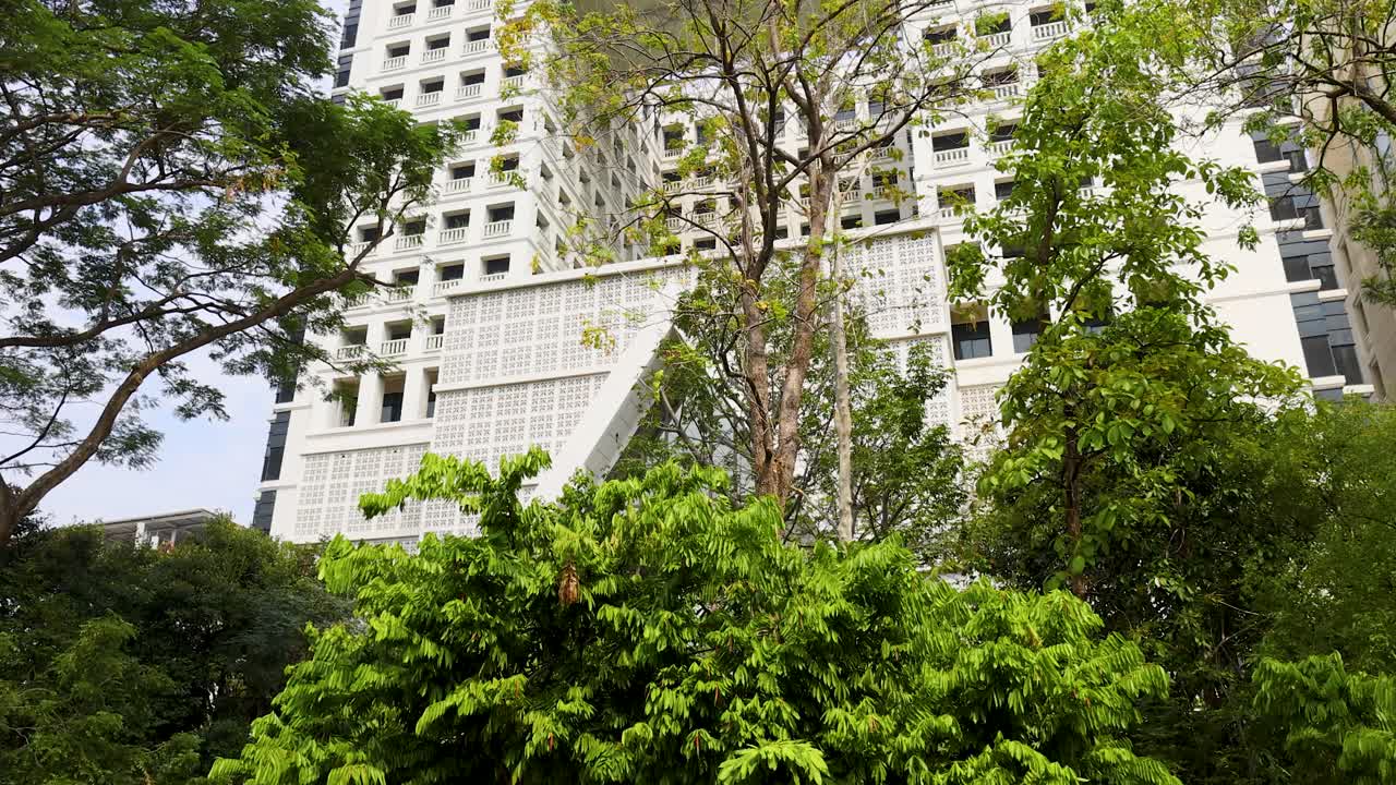 Uma inclinação ascendente lenta revela um edifício contemporâneo branco de vários andares atrás de exuberantes árvores tropicais verdes