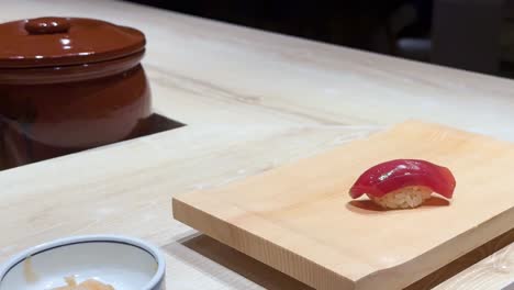 a piece of sushi is picked up with chopsticks from a wooden board, next to a ceramic bowl.