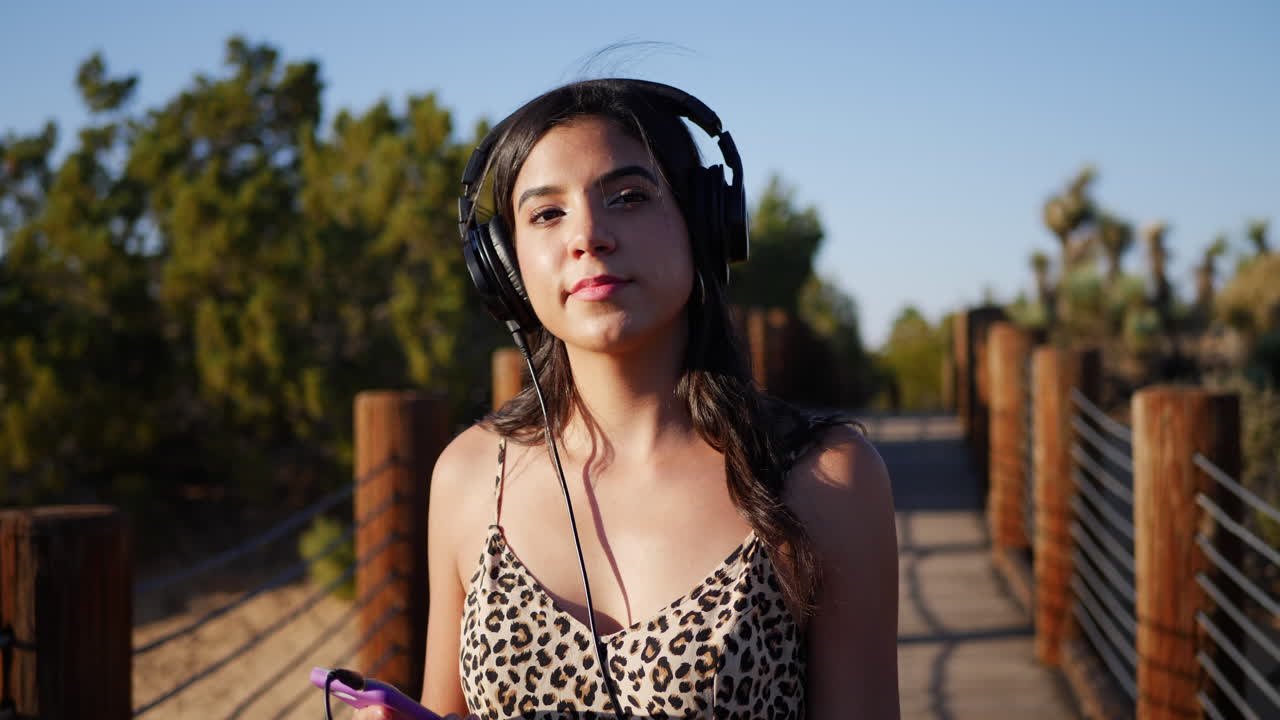 una linda joven hispana escuchando música pop en sus auriculares y smartphone mientras camina en cámara lenta al aire libre bajo la luz del sol