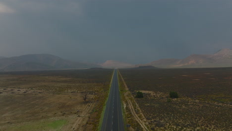 Backwards-fly-above-empty-straight-road-in-barren-landscape.-Mountain-ridge-looms-in-background.-South-Africa