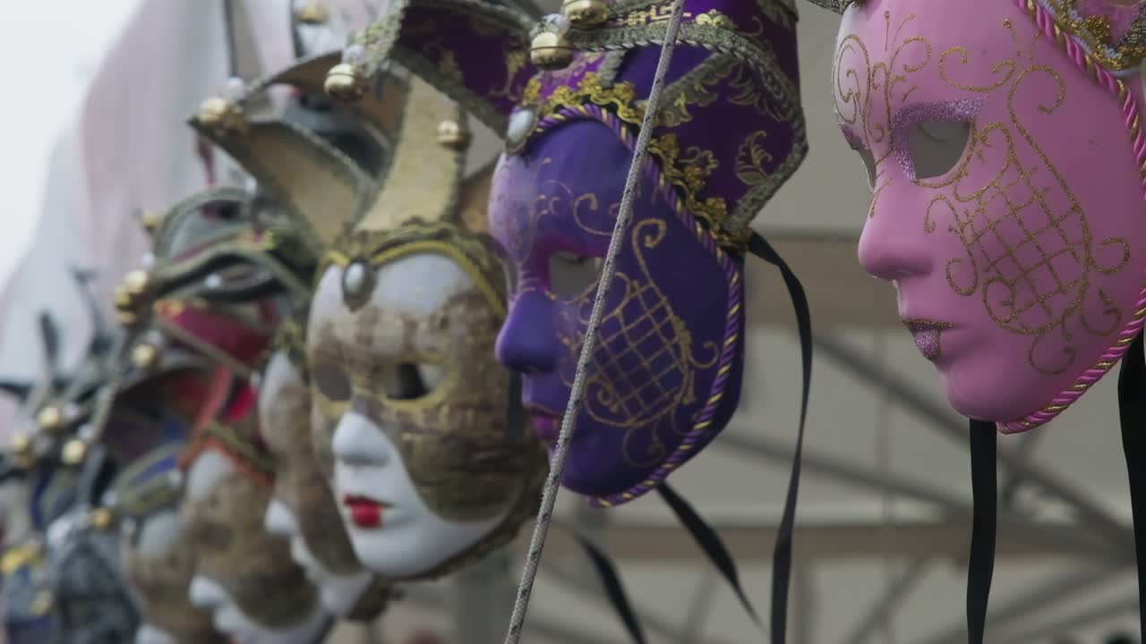 Maschere di carnevale veneziane ornate appese e che oscillano lentamente in una bancarella del mercato. Pezzi di costume decorativi viola e rosa con intricati motivi dorati esposti per la vendita.