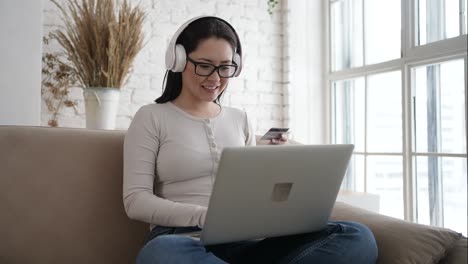 happy young asian woman doing payments online or shopping in internet store holding credit card using laptop computer