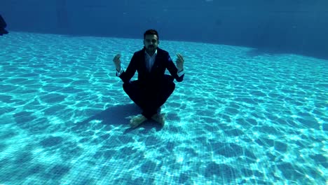 a man in a suit and a white shirt meditates under the water in the pool in a lotus position. he sits on the bottom and looks around. slow motion. action camera underwater.