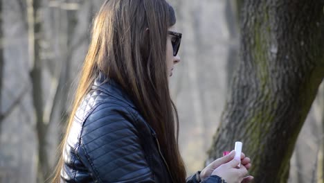 Beautiful-young-woman-sitting-on-a-bench-in-the-public-park-and-putting-on-red-lipstick-while-the-sun-shines-on-her-back