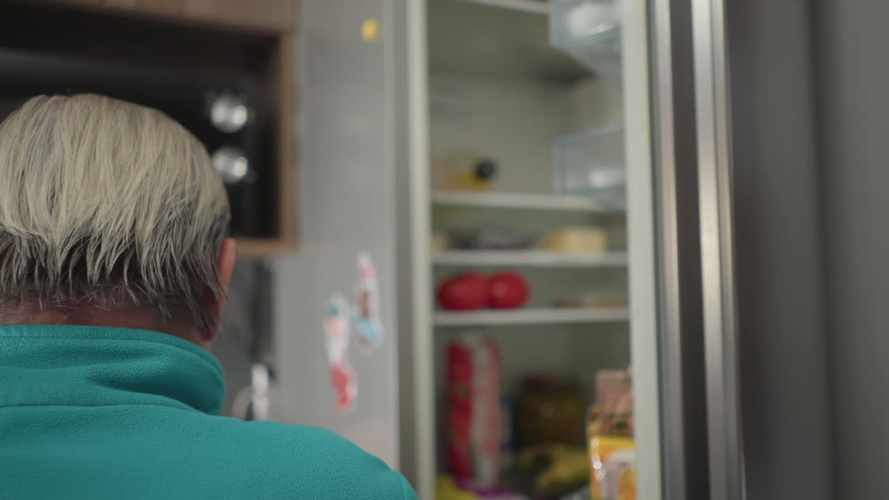 Elderly woman in green sweatshirt reaching to grab items from top shelf of refrigerator. Focus on daily living routine in a well-organized kitchen, showing independence and healthy lifestyle