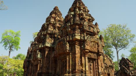 ancient banteay srei temple with intricate carvings, warm lighting, and lush trees under a perfect blue sky. honey coloured historic 10th century khmer remains in jungle setting in cambodia.